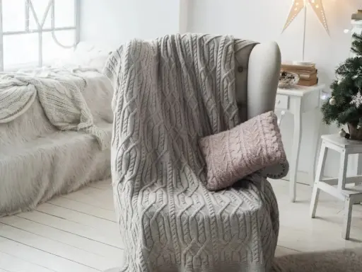 Manta de punto gris con trenzado, estilo plaid Bassetti, sobre sillón junto a árbol de Navidad.