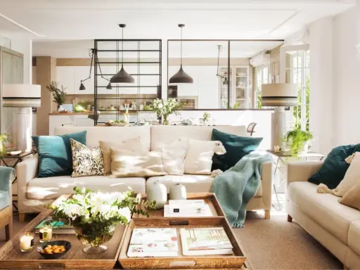 Salones con color: sofás beige, cojines turquesas y mantas suaves crean un ambiente acogedor. Una mesa de centro de madera con flores y libros completa la escena.
