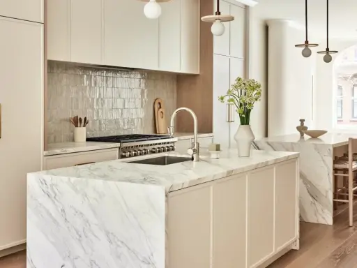 Elegante cocina atemporal con isla de mármol, gabinetes crema y electrodomésticos de acero inoxidable. Un jarrón con flores ilumina el espacio.