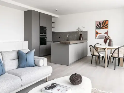 Salón moderno y cocina integrada, ideal para amueblar piso de alquiler. Sofá gris, mesa redonda con sillas de ratán y arte abstracto.