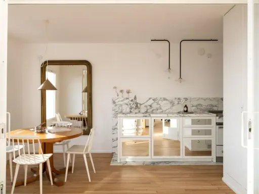 Comedores pequeños con encanto, con mesa redonda de madera, sillas blancas y un gran espejo dorado. La cocina integrada tiene encimera de mármol y armarios con puertas de espejo.