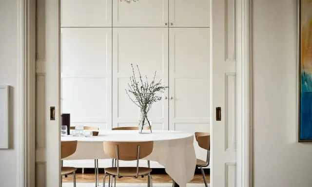 Comedor con mesa cubierta, sillas de madera y una pared en blanco RAL 9010. Un jarrón con ramas decora la mesa.