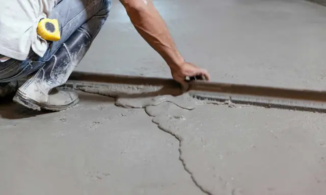 Obrero con botas de goma y cinta métrica en el bolsillo, usando una regla para nivelar suelo de cemento fresco.