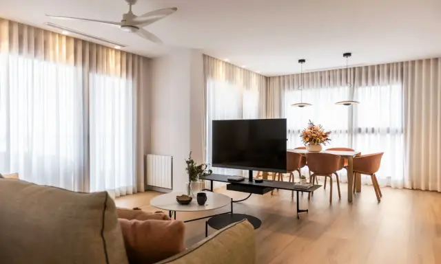 Acogedor salón de 20 metros cuadrados con sofá, mesa de centro, TV, y comedor. Cortinas blancas y suelo de madera crean un ambiente luminoso.