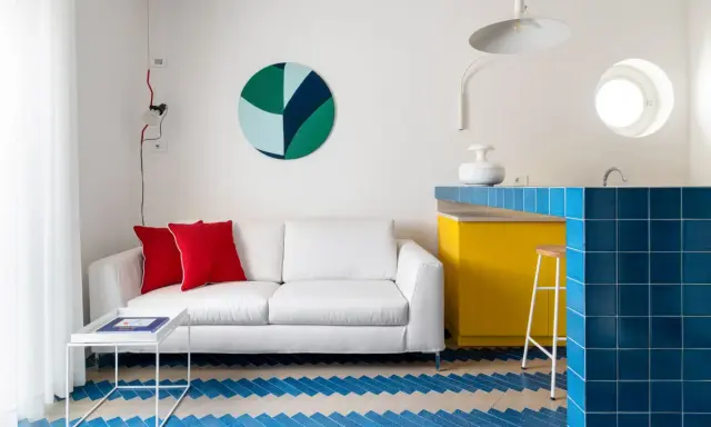 Sofá blanco con cojines rojos, mesa auxiliar blanca y suelo con diseño de espiga en colores claros y azul. Barra de cocina amarilla y azul.