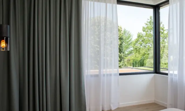 Cortinas bandalux grises y blancas en un dormitorio moderno, con vistas a un jardín frondoso.