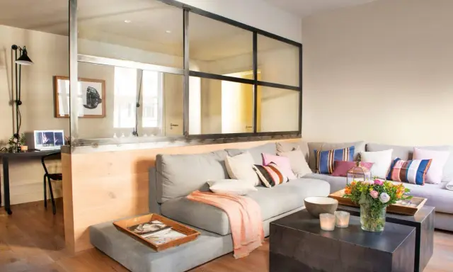 Un salón moderno con una pared de cristal que separa un área de trabajo. Un sofá gris con cojines coloridos y una manta rosa.