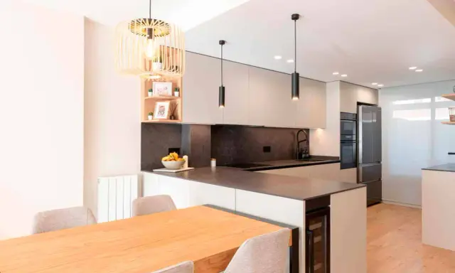 Moderna cocina en L con isla, electrodomésticos integrados, encimera de cuarzo y mesa de madera.