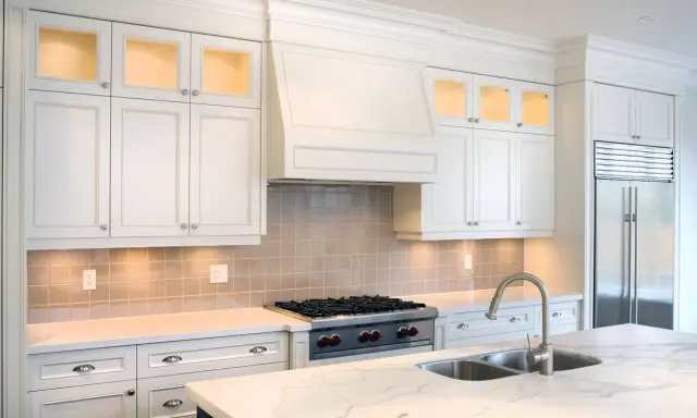 Cocina moderna con campana extractora decorativa sobre estufa. Gabinetes blancos, encimera de mármol y refrigerador de acero inoxidable.