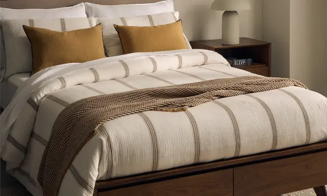 Cama con ropa de cama a rayas, cojines mostaza y manta tejida. Las bassols sabanas aportan un toque acogedor.