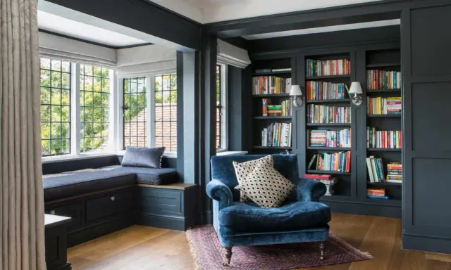 Acogedor rincón lectura salón con sillón azul, ventana con banco y estanterías llenas de libros.