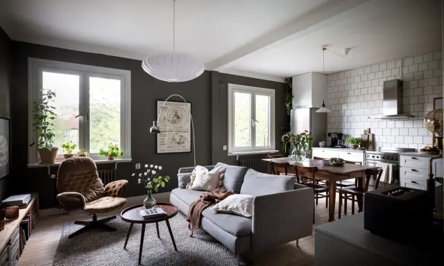 Un salón rectangular amueblado con sofá gris, sillón marrón, mesa de centro y alfombra. Cocina abierta con mesa de comedor.