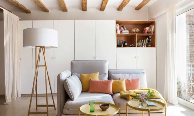 Ideas para decorar piso alquiler low cost: sofá gris, cojines coloridos, mesa auxiliar dorada, lámpara de pie y estantería con libros.