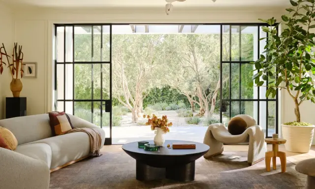 Salones elegantes con sofás curvos, una silla acogedora y grandes ventanales que conectan con un jardín de olivos.