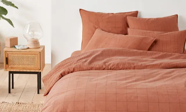 Cama con funda nordica terracota a cuadros y rayas. Mesita de noche de madera con lámpara y planta.