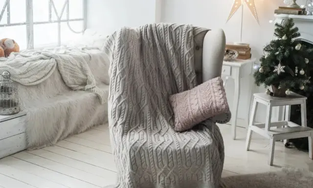 Manta de punto gris con trenzado, estilo plaid Bassetti, sobre sillón junto a árbol de Navidad.