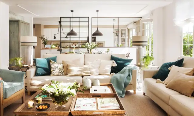 Salones con color: sofás beige, cojines turquesas y mantas suaves crean un ambiente acogedor. Una mesa de centro de madera con flores y libros completa la escena.