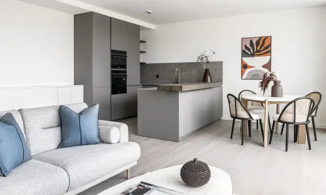 Salón moderno y cocina integrada, ideal para amueblar piso de alquiler. Sofá gris, mesa redonda con sillas de ratán y arte abstracto.