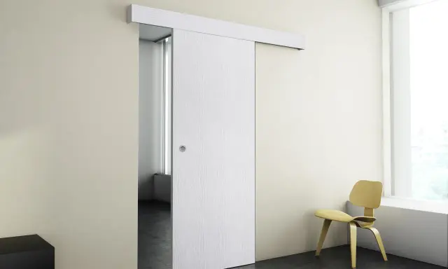 Puerta corredera blanca moderna instalada en pared beige, con silla amarilla y ventana al fondo.