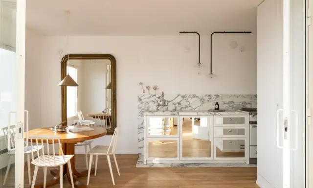 Comedores pequeños con encanto, con mesa redonda de madera, sillas blancas y un gran espejo dorado. La cocina integrada tiene encimera de mármol y armarios con puertas de espejo.
