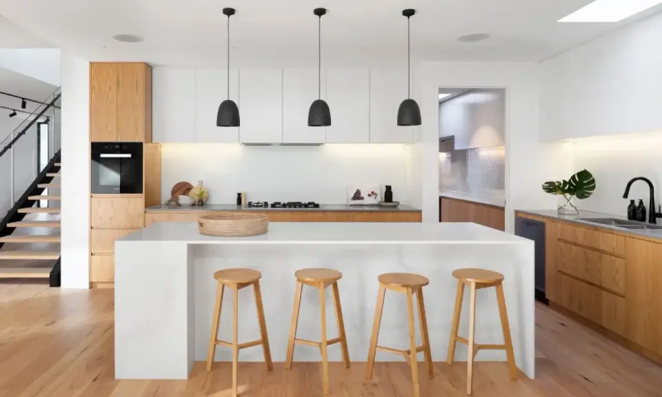 Cocinas modernas con isla blanca, taburetes de madera y gabinetes de roble. Escalera de diseño minimalista al fondo.