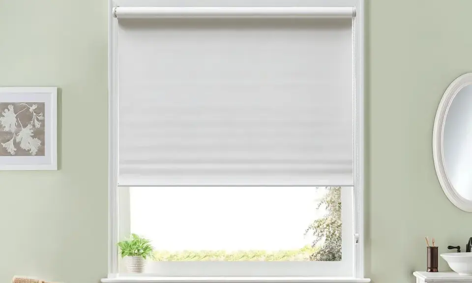 Persiana enrollable blanca que cubre la mayor parte de la ventana. Un pequeño paquete de toallas verdes y blancas descansa en la repisa.