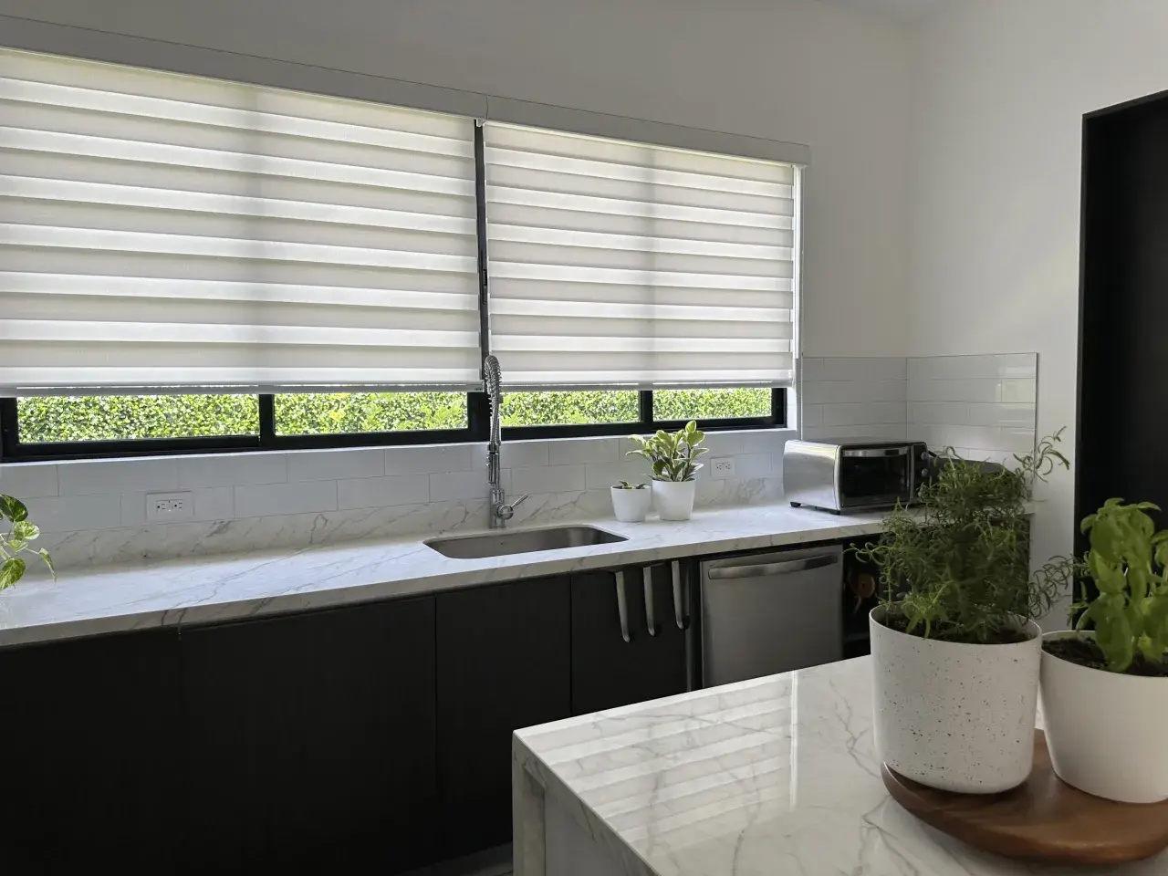 Cocina moderna con encimeras de mármol, electrodomésticos de acero inoxidable y dos tipos de persianas que filtran la luz natural.