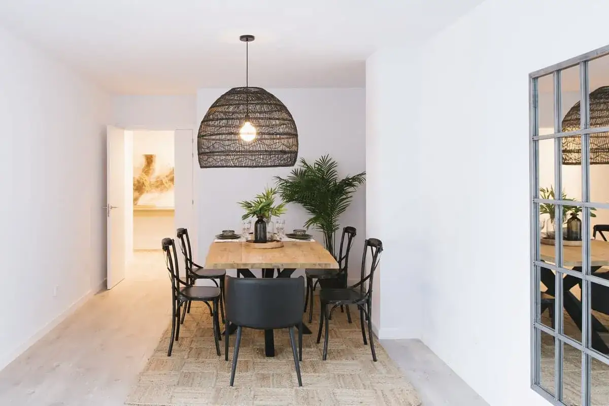 Mesa de comedor de madera r&uacute;stica con sillas negras, planta y un gran colgante de mimbre. Ideal para poner encima de una mesa de comedor.