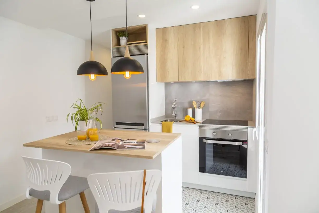 Cocinas abiertas al salón pequeñas con barra de desayuno, electrodomésticos modernos y dos taburetes blancos.