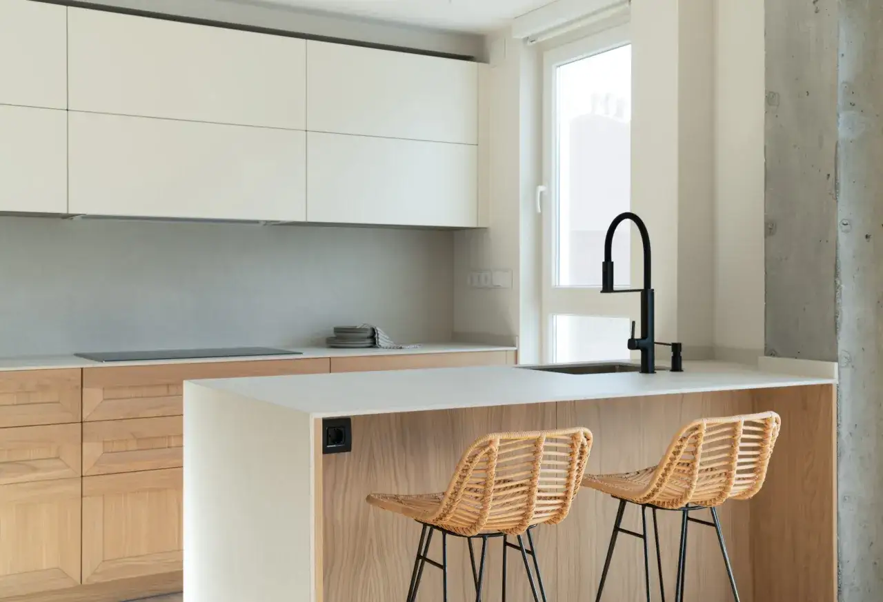 Cocinas modernas con isla central, taburetes de mimbre y grifería negra. Muebles de madera y encimera clara.