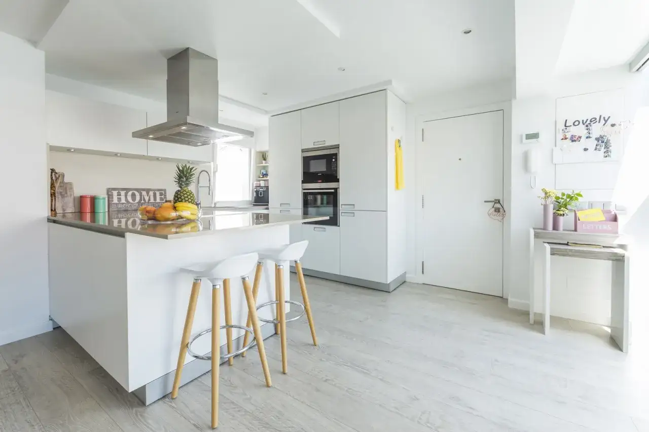 Cocina moderna con isla central, taburetes blancos y suelos para cocinas blancas de madera clara.