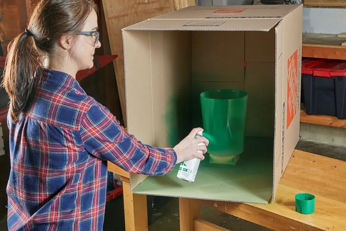Mujer pintando un jarr&oacute;n verde con pintura en spray dentro de una caja de cart&oacute;n. Usa pintura para pintar pl&aacute;stico.