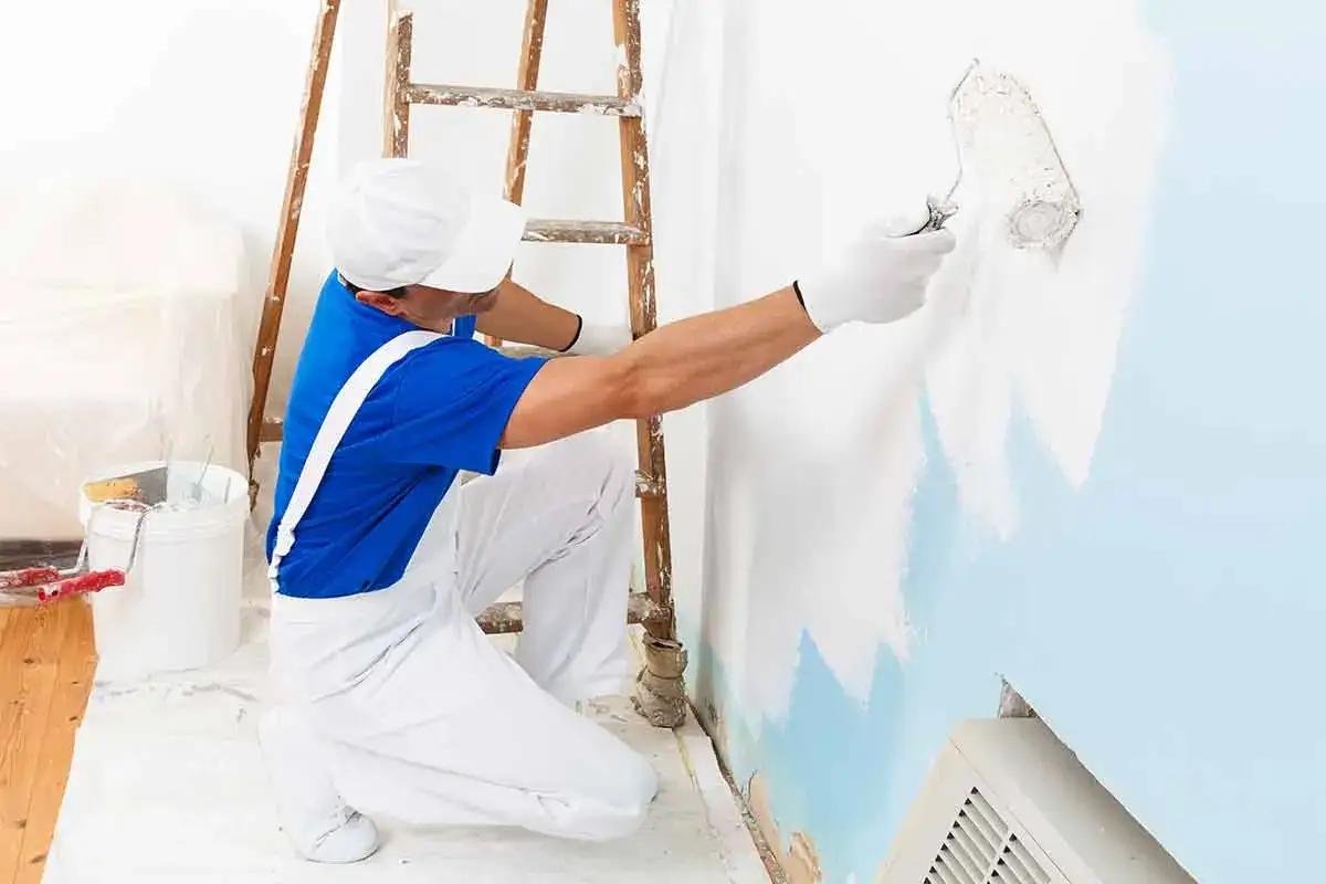 Pintor con rodillo aplica pintura al agua en pared azul claro. Escalera de madera y cubeta de pintura a un lado.