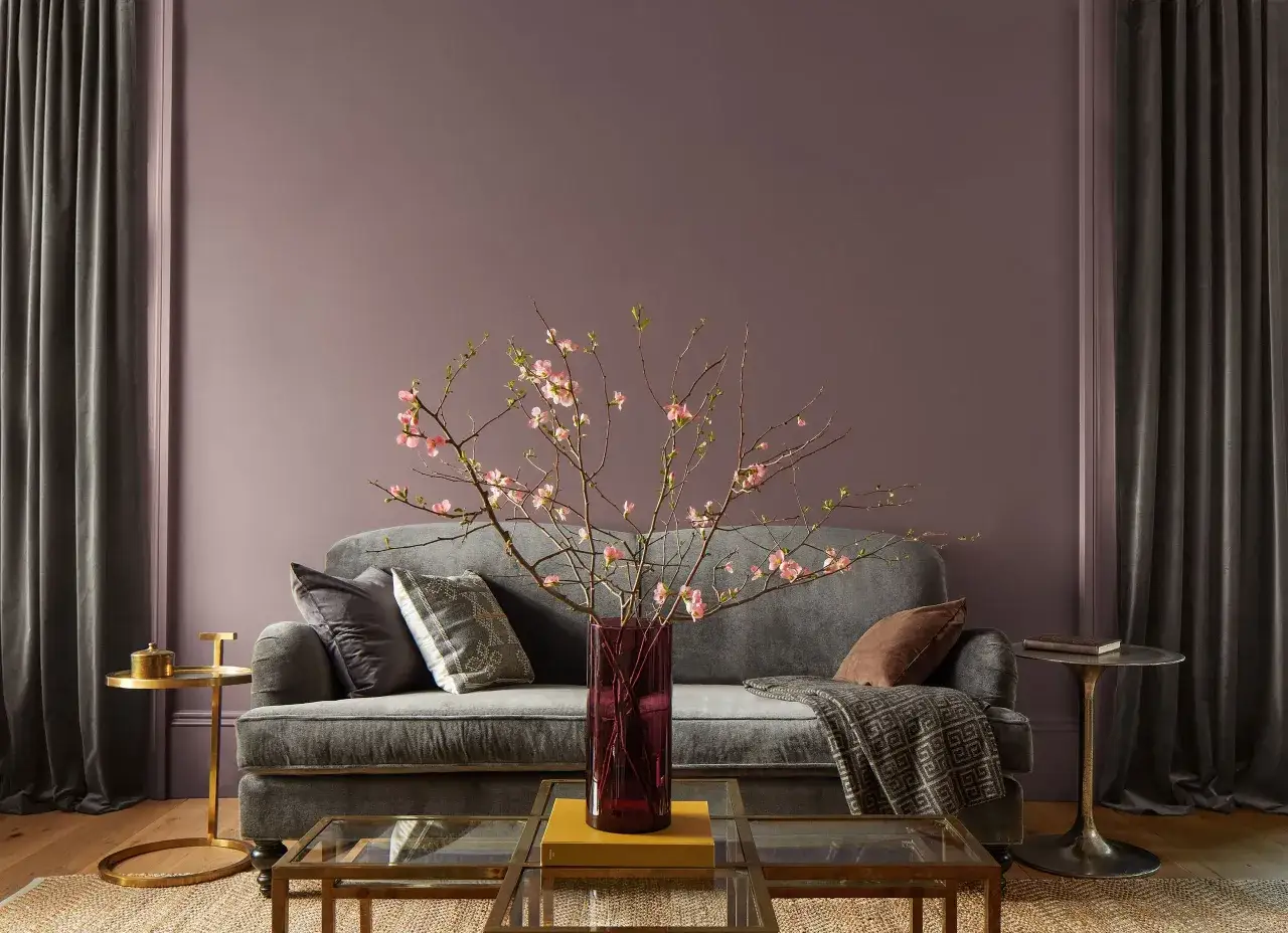 Sof&aacute; gris con cojines y manta, sobre mesas de cristal y lat&oacute;n. Flores rosadas en jarr&oacute;n morado. Paredes en tonos malva, uno de los colores de moda para pintar casa.