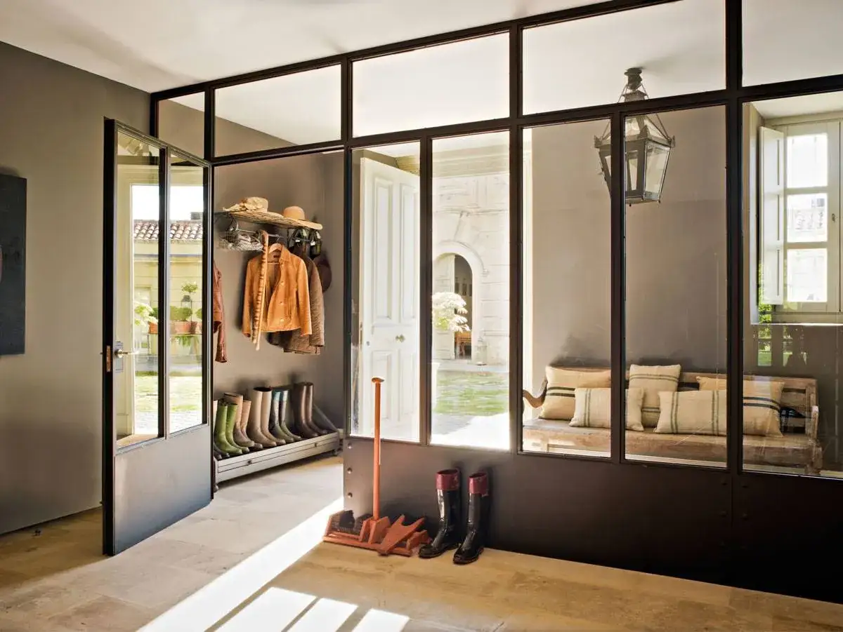Entrada acogedora con una pared de cristal que da a un patio. Hay botas, chaquetas y un banco con cojines.