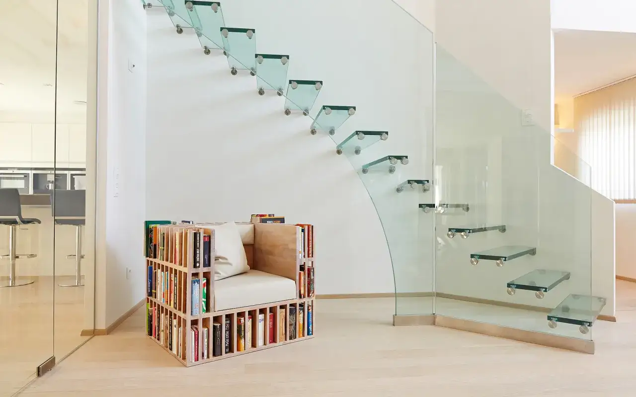 Modernas escaleras vistas en el sal&oacute;n, con pelda&ntilde;os de cristal flotantes y una barandilla de vidrio curvo. Un sill&oacute;n integrado en una estanter&iacute;a de libros completa el espacio.