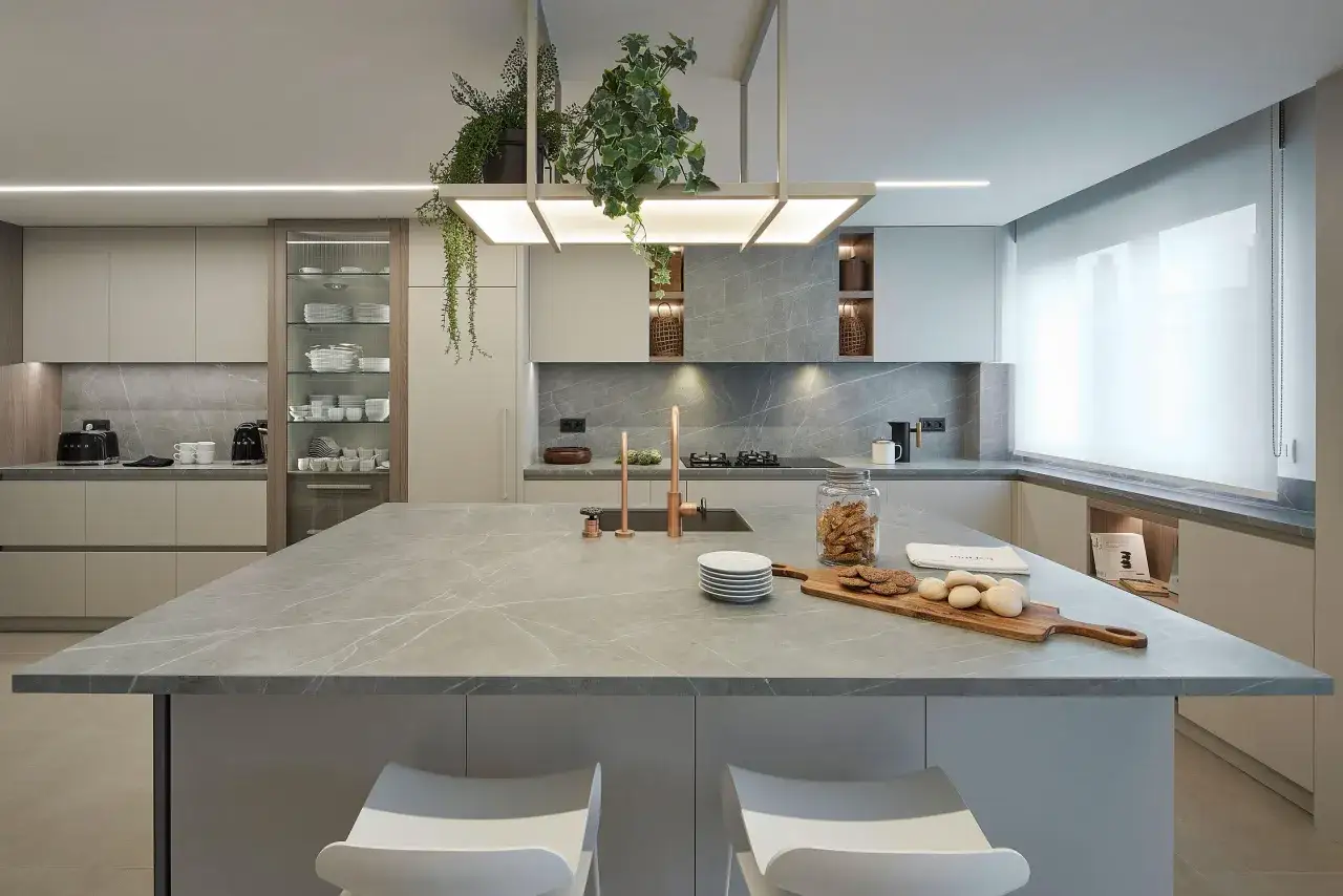 Moderna cocina con isla central de m&aacute;rmol gris. Estanter&iacute;as abiertas con plantas colgantes y armarios minimalistas.