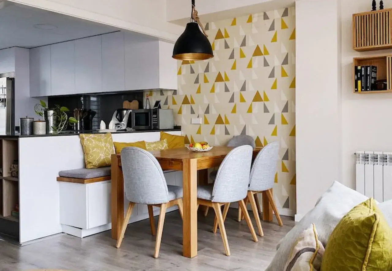 Comedor moderno con cocinas con papel pintado de tri&aacute;ngulos amarillos y grises. Mesa de madera, sillas grises y banco con cojines.