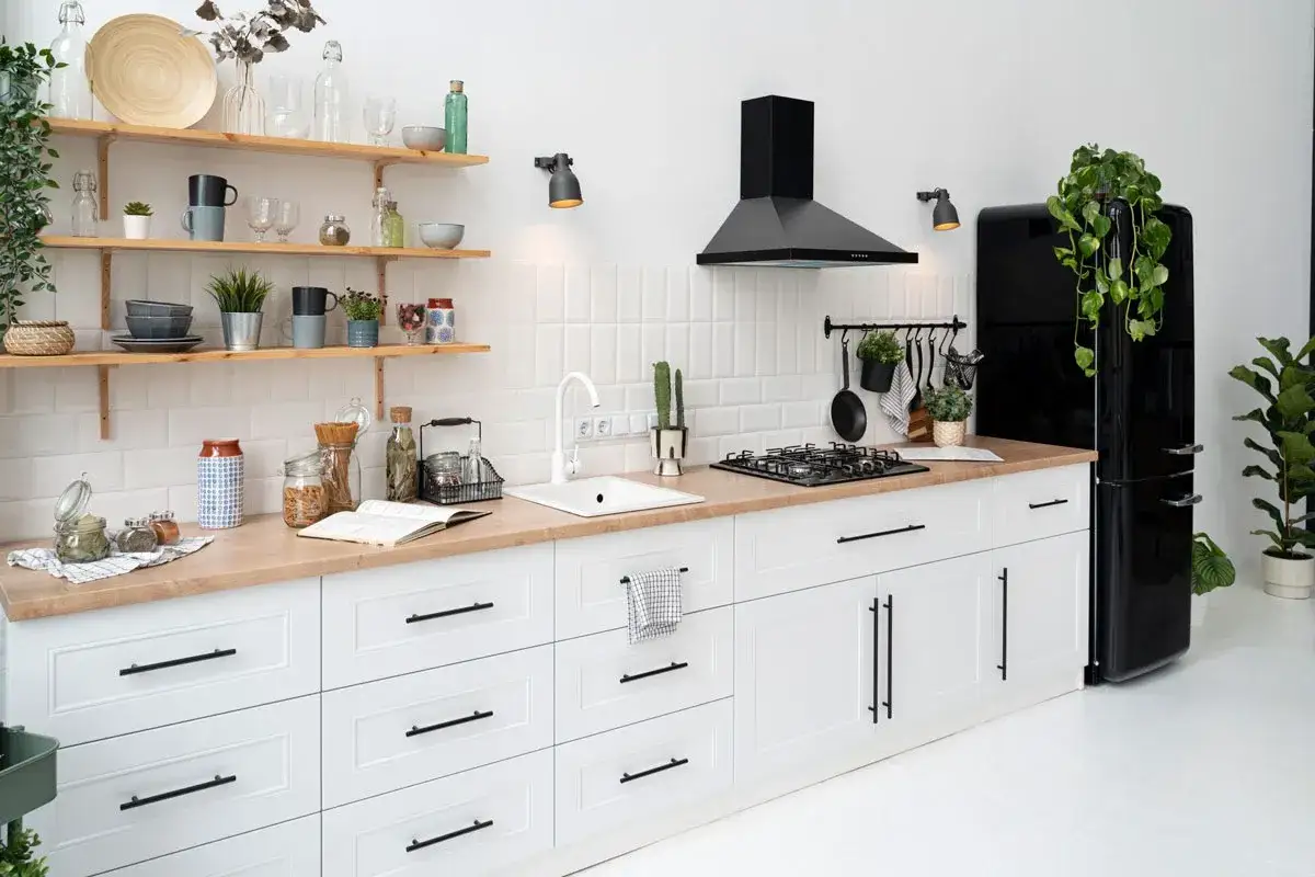Cocina blanca y luminosa con encimera de madera, ideal para reformar cocina sin obras. Estanterías con menaje, electrodomésticos negros y plantas verdes.