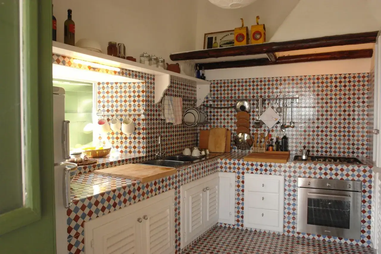 Encantadora cocina de obra con azulejos de rombos en tonos tierra y azul. Utensilios de cocina colgados y una encimera de madera completan el ambiente r&uacute;stico.