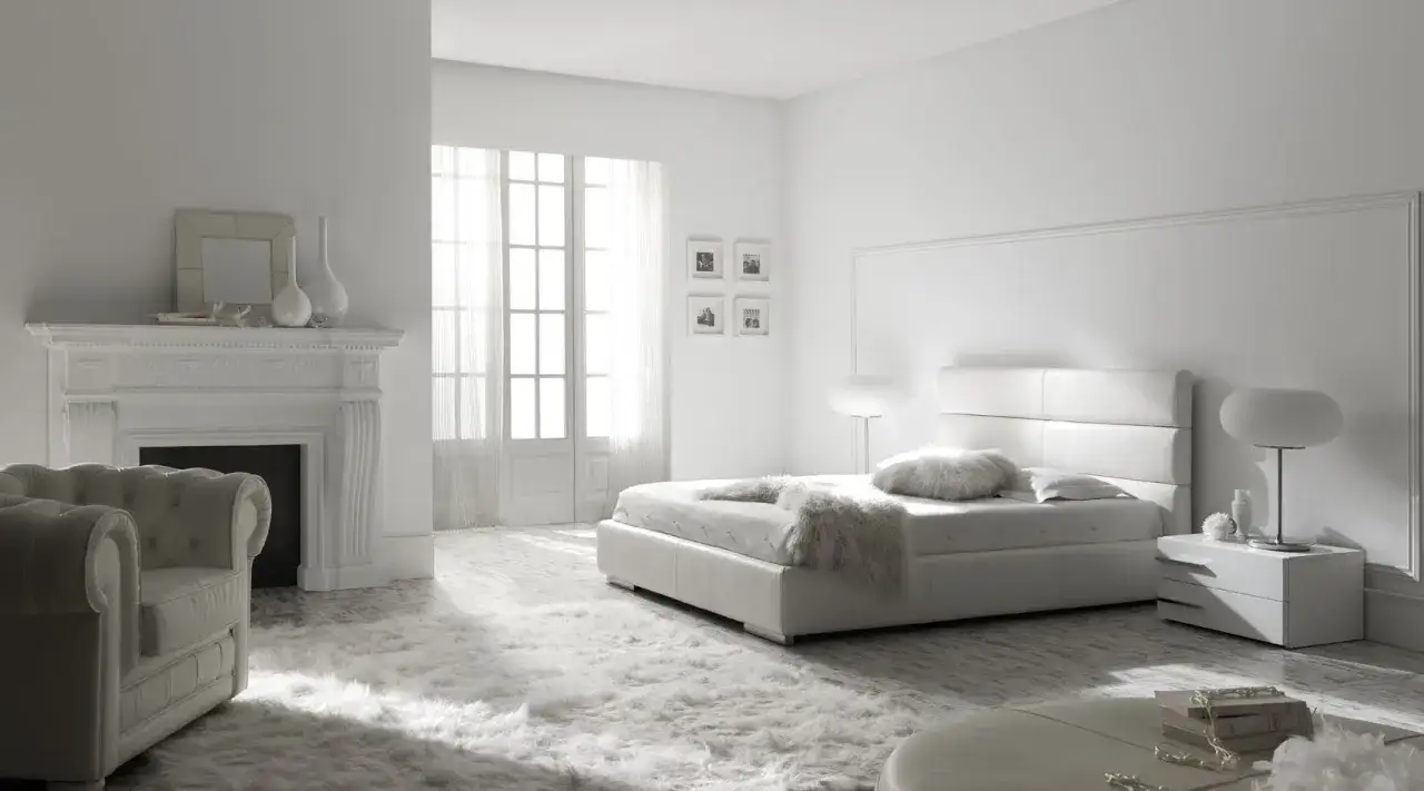 Dormitorio minimalista con cama blanca, chimenea y sillón, todo en colores claros que transmiten serenidad.