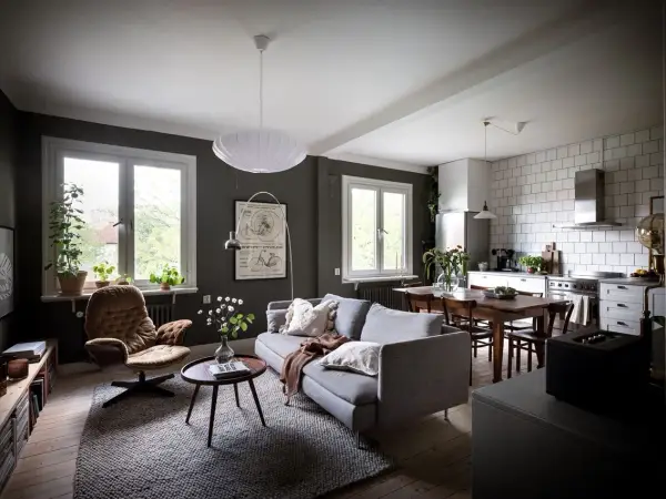 Un salón rectangular amueblado con sofá gris, sillón marrón, mesa de centro y alfombra. Cocina abierta con mesa de comedor.