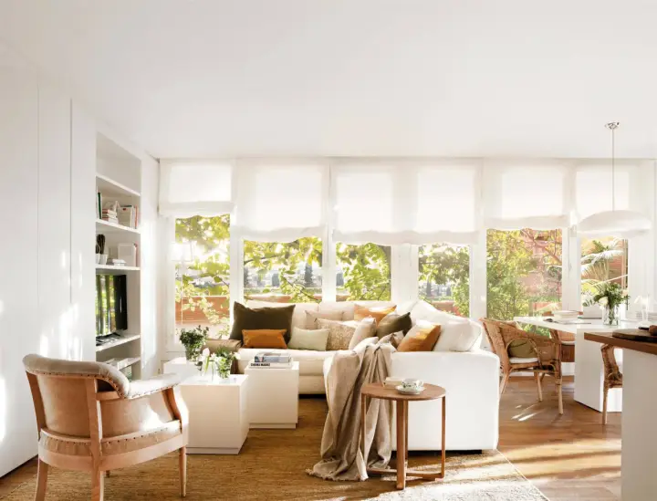 Acogedor salón comedor dos ambientes con sofá blanco, sillón de lino y mesa de mimbre. Luz natural entra por grandes ventanales.