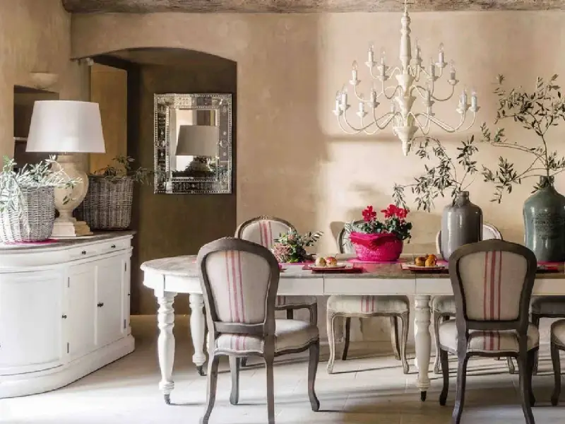 Comedor con estilo provenzal moderno. Mesa con sillas tapizadas, lámpara de araña, jarrón con ramas y flores. Aparador blanco a la izquierda.